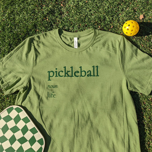 A pickleball shirt - a shirt in pickle green color, in emerald ink a pickleball definition reads "Pickleball, noun, life"
